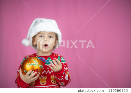Cute child in a festive sweater and Santa hat holds a Christmas decoration on a pink background. The child is happy about the New Year and Christmas. Suitable for holiday projects. 119560533
