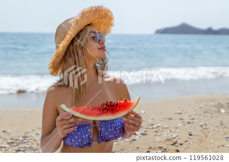 Portrait of young beautiful smiling blond woman in swimsuit wearing sunglasses and hat while happily sitting with slice of watermelon on beach Portrait of young beautiful smiling blond woman in swimsuit wearing sunglasses and hat while happily sitting with slice of watermelon on beach 119561028