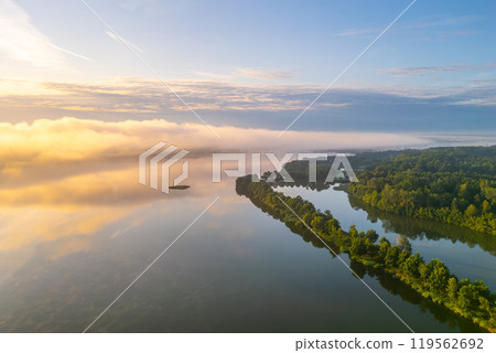 A tranquil morning at Rozmberk Pond reveals mist rising from the calm water. Lush greenery lines the shore, reflecting soft hues of dawn, creating a serene backdrop for nature's beauty. 119562692