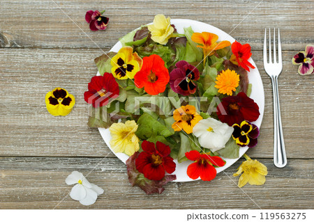 Table top view of the plate with salad made of edible flowers (nasturtium, pansy, calendula) and lettuce on the wooden table with a fork. 119563275