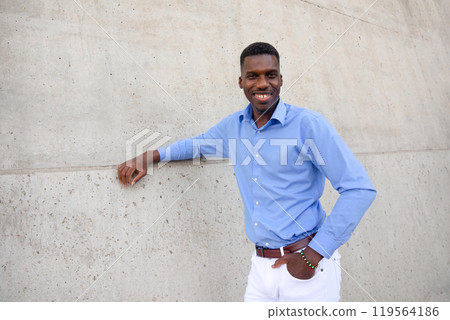 Portrait Of Handsome Smiling With Teeth African American Man wearing White Jeans and Blue Shirt, Brown Belt Outdoor, Concrete Wall on Background. Copy Space Fashionable Male, Ivorian. Horizontal 119564186