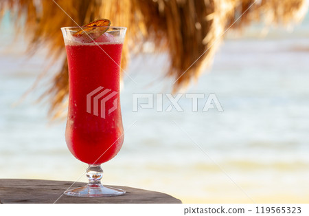 Cold red strawberry cocktail in a glass with a dry orange slice on the wooden table on the beach under the straw roof umbrella, the beach and the sea is on the background. Cold red strawberry cocktail in a glass with a dry orange slice on the wooden table on the beach under the straw roof umbrella, the beach and the sea is on the background. 119565323