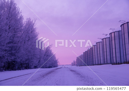 Picturesque winter sunrise in the grain farm in prairies. Granaries along the country road in purple and pink morning light. Picturesque winter sunrise in the grain farm in prairies. Granaries along the country road in purple and pink morning light. 119565477