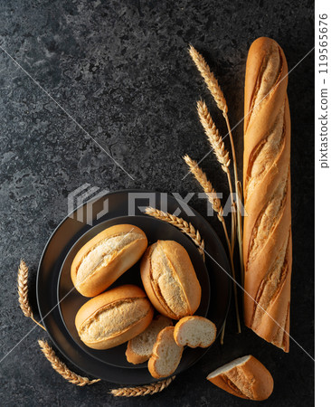 Baguette, French buns, and wheat ears on a black stone table. 119565676