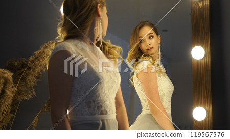 Bride admires herself in a white lace dress while posing in front of a mirror Bride admires herself in a white lace dress while posing in front of a mirror 119567576