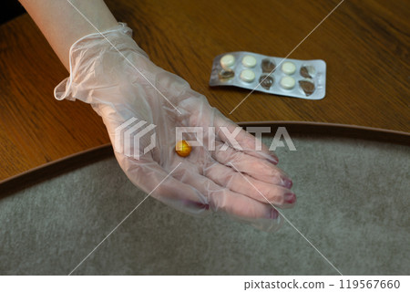 Hand in medical glove holds expired tablet brown color against background of plate of tablets that have lost their expiration date, monitoring suitability and freshness of medicaments 119567660