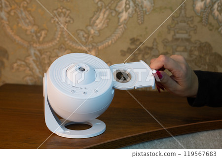 Female hand inserts galaxy image cartridge into compact home projector for space exploration, scientific device for home use, room projector on shelf 119567683