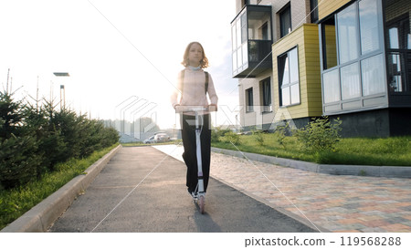 Cheerful schoolgirl rides her scooter down a sunlit street as she heads to school, enjoying the bright morning 119568288