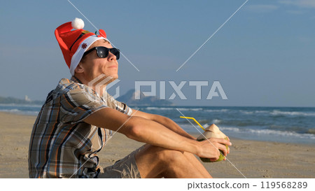 Man in a festive Christmas hat and sunglasses enjoys coconut water while lounging on a tropical beach during the winter holidays 119568289