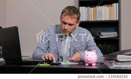 Focused man organizes his finances at his desk, counting coins and depositing them into a pink piggy bank Focused man organizes his finances at his desk, counting coins and depositing them into a pink piggy bank 119568292
