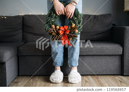Person holding handcrafted Christmas wreath with pinecones and red bow, standing in cozy living room. DIY holiday traditions, handmade decor, and personal creativity. Person holding handcrafted Christmas wreath with pinecones and red bow, standing in cozy living room. DIY holiday traditions, handmade decor, and personal creativity. 119568957