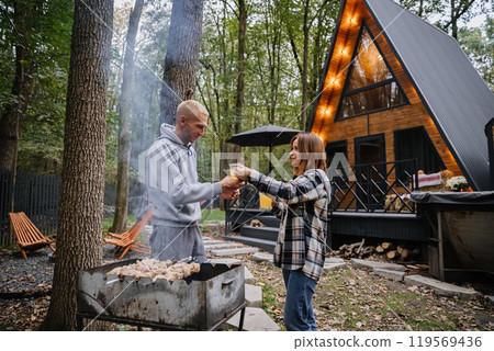 Couple toasting with beer near grill in a cozy outdoor setting, enjoying barbecue and nature retreat 119569436