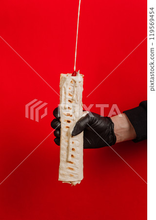 Dripping sauce on wrap held by gloved hand, highlighting street food presentation on red backdrop 119569454