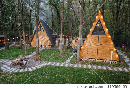 Modern wooden A-frame cabins in forest with outdoor seating and fire pit area, relaxation time Modern wooden A-frame cabins in forest with outdoor seating and fire pit area, relaxation time 119569463