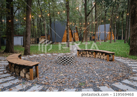Cozy fire pit seating area in forest glamping with glowing string lights and modern wooden cabins Cozy fire pit seating area in forest glamping with glowing string lights and modern wooden cabins 119569468