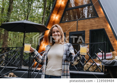 Smiling woman holding beer outdoors near cozy cabin, enjoying a relaxing autumn getaway in nature 119569484