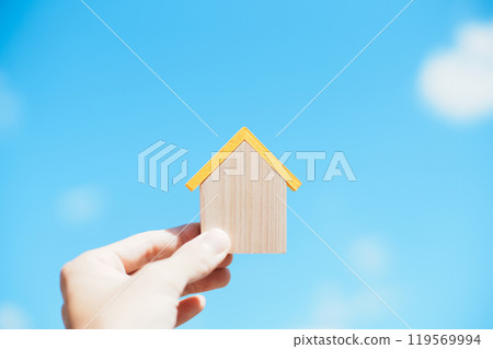 Person's hand holding a small wooden house with blue sky background 119569994
