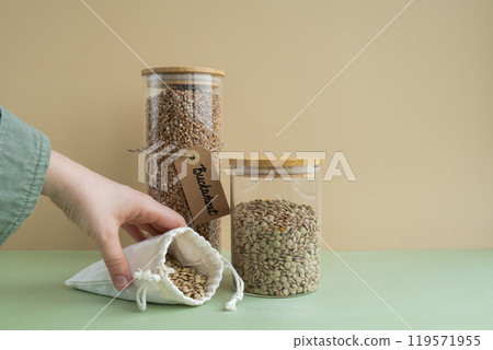Unrecognizable woman refill Refillable glass jar with raw buckwheat and green lentil porridge in cotton bag. Product format eco-friendly living. Filling reusable container Whole spelt grain species Unrecognizable woman refill Refillable glass jar with raw buckwheat and green lentil porridge in cotton bag. Product format eco-friendly living. Filling reusable container Whole spelt grain species 119571955