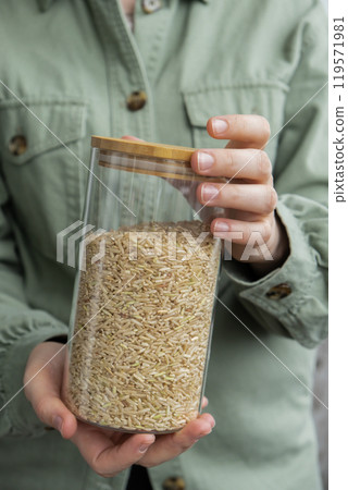 Unrecognizable woman holding refillable glass jar with raw brown rice porridge. Refill product format eco-friendly living. Filling reusable container Whole spelt grain species. High protein fiber-rich Unrecognizable woman holding refillable glass jar with raw brown rice porridge. Refill product format eco-friendly living. Filling reusable container Whole spelt grain species. High protein fiber-rich 119571981