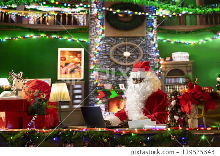Santa Claus searches online for gift locations using his laptop in a festively decorated cabin at the North Pole. Saint Nicholas is connected to the world, making Christmas magic happen. 119575343