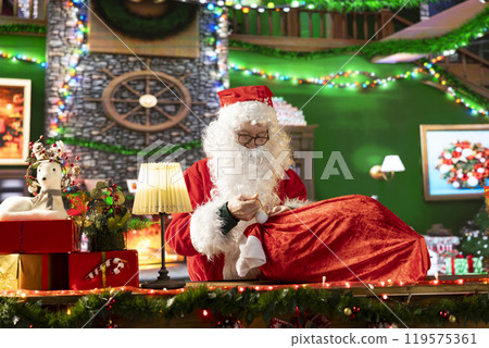 White bearded old man in costume tying his sack full of gifts, preparing for the journey around the world. Spreading the winter holiday spirit and surprising children, x mas tradition. White bearded old man in costume tying his sack full of gifts, preparing for the journey around the world. Spreading the winter holiday spirit and surprising children, x mas tradition. 119575361