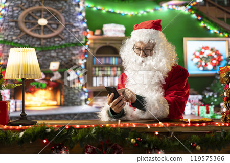Father Christmas uses a mobile phone app in his North Pole cabin, browsing the web to ensure toys and gifts are ready for delivery on winter holiday eve. Festive season full of magic. Father Christmas uses a mobile phone app in his North Pole cabin, browsing the web to ensure toys and gifts are ready for delivery on winter holiday eve. Festive season full of magic. 119575366