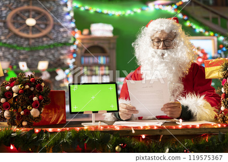 Father Christmas looking at his naughty or nice list next to isolated copy space screen, making preparations for x mas journey to bring gifts. Santa Claus reading wishes from children. Father Christmas looking at his naughty or nice list next to isolated copy space screen, making preparations for x mas journey to bring gifts. Santa Claus reading wishes from children. 119575367