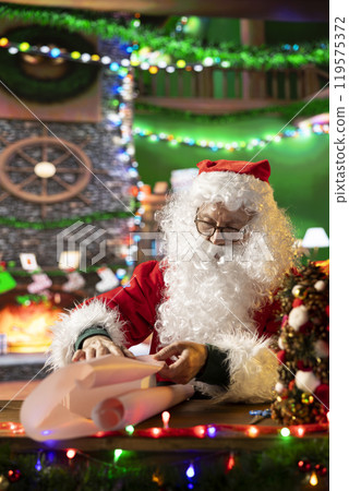 Santa Claus wraps a festive gift box by the fire in his North Pole workshop, surrounded by holiday lights and decorations. Father Christmas prepares gifts and surprises for winter celebration. 119575372