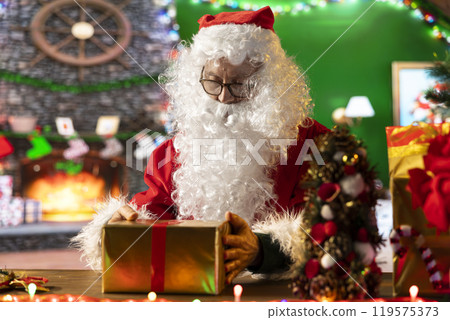 Santa Claus wraps a festive gift box by the fire in his North Pole workshop, surrounded by holiday lights and decorations. Father Christmas prepares gifts and surprises for winter celebration. 119575373