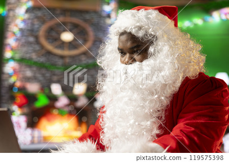 Santa Claus uses a laptop in his festive log cabin navigating online webpages to track gifts. The North Pole is filled with Christmas magic as Saint Nicholas embraces the digital age. Santa Claus uses a laptop in his festive log cabin navigating online webpages to track gifts. The North Pole is filled with Christmas magic as Saint Nicholas embraces the digital age. 119575398