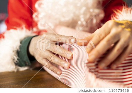 Saint Nicholas wraps a festive gift box surrounded by Christmas lights and ornaments in his workshop. Santa prepares for the holiday season, packing presents to bring joy to children. Close up. Saint Nicholas wraps a festive gift box surrounded by Christmas lights and ornaments in his workshop. Santa prepares for the holiday season, packing presents to bring joy to children. Close up. 119575436