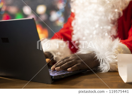 Father Christmas is browsing websites on his laptop and checking holiday gift lists from his North Pole cabin. The festive season is alive with online magic and holiday traditions. 119575441