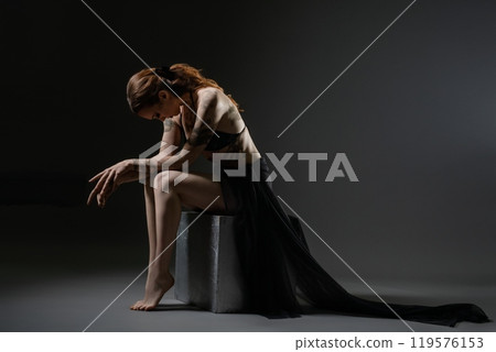 A contemplative dancer in a flowing dress poses on a stool in dim lighting 119576153