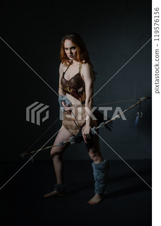 A warrior woman poses with a bow and arrow in a dramatic dark setting A warrior woman poses with a bow and arrow in a dramatic dark setting 119576156
