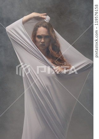 Female model draped in sheer fabric poses dramatically in a misty studio setting Female model draped in sheer fabric poses dramatically in a misty studio setting 119576158