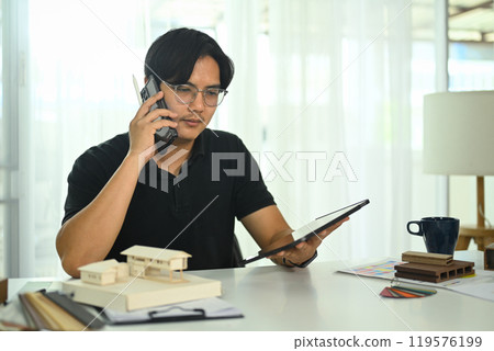 Asian male talking on mobile phone while working on architectural plans in the office 119576199