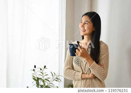 Smiling young woman in a cozy sweater enjoying a hot drink and gazing out the window Smiling young woman in a cozy sweater enjoying a hot drink and gazing out the window 119576231