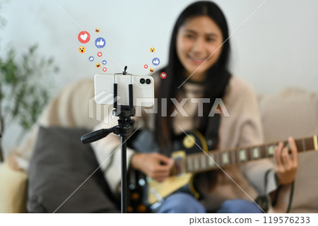 Young woman playing guitar during live streaming on smartphone at home Young woman playing guitar during live streaming on smartphone at home 119576233
