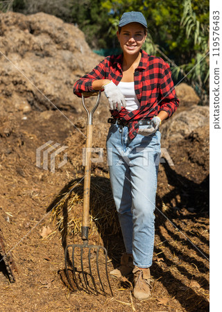 Young woman with rake of pile of manure Young woman with rake of pile of manure 119576483