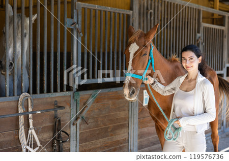 Young woman leads horse out of stable 119576736