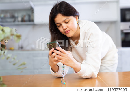 Young brunette browsing through websites on smartphone in home kitchen Young brunette browsing through websites on smartphone in home kitchen 119577005