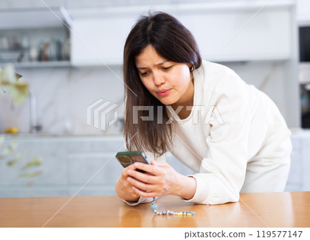 Upset young woman standing with smartphone in home kitchen Upset young woman standing with smartphone in home kitchen 119577147