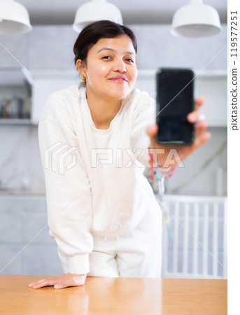 Cheerful young woman showing phone in home kitchen Cheerful young woman showing phone in home kitchen 119577251