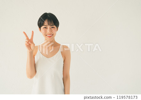 A young woman in a camisole makes a gesture of 2 in front of a white background 119578173