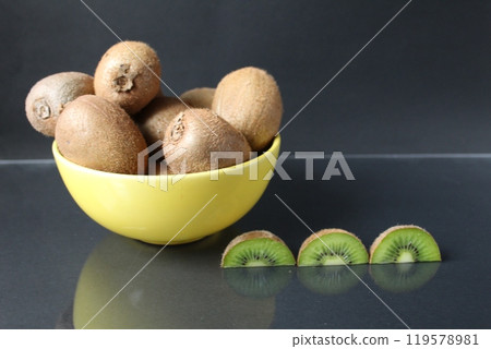 still life of kiwi fruit in a yellow bowl plate and pieces of mug next to on a black background with a reflection of there is a copyspace 119578981