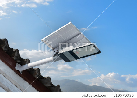 Mini solar cell panel and hd floodlight panal installed together under house eaves to store and use the power from the sunlight with street light and bulbs at night, sunset background, soft focus. Mini solar cell panel and hd floodlight panal installed together under house eaves to store and use the power from the sunlight with street light and bulbs at night, sunset background, soft focus. 119579356