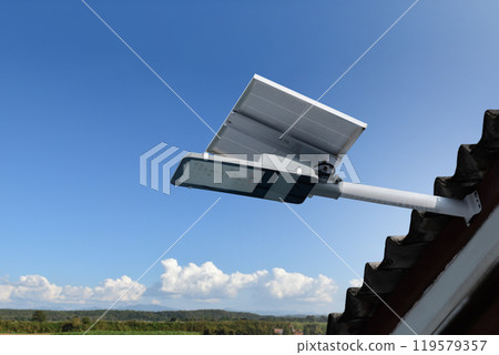 Mini solar cell panel and hd floodlight panal installed together under house eaves to store and use the power from the sunlight with street light and bulbs at night, sunset background, soft focus. Mini solar cell panel and hd floodlight panal installed together under house eaves to store and use the power from the sunlight with street light and bulbs at night, sunset background, soft focus. 119579357