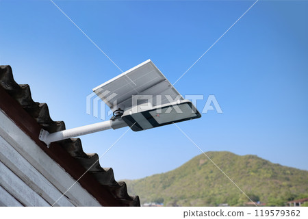 Mini solar cell panel and hd floodlight panal installed together under house eaves to store and use the power from the sunlight with street light and bulbs at night, sunset background, soft focus. 119579362