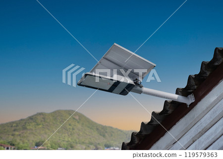 Mini solar cell panel and hd floodlight panal installed together under house eaves to store and use the power from the sunlight with street light and bulbs at night, sunset background, soft focus. 119579363