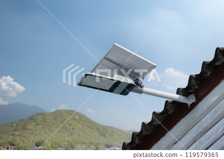 Mini solar cell panel and hd floodlight panal installed together under house eaves to store and use the power from the sunlight with street light and bulbs at night, sunset background, soft focus. 119579365